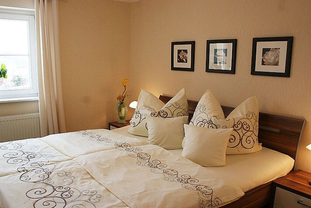 A neatly made bed with a patterned bedspread and decorative pillows, a bedside table with a vase, and three framed pictures on the wall.
