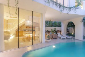 A modern indoor pool area with a glass-enclosed kitchen and dining area, lounge chairs, and a ceiling fan. - Kona Resort Pererenan E (Badung)
