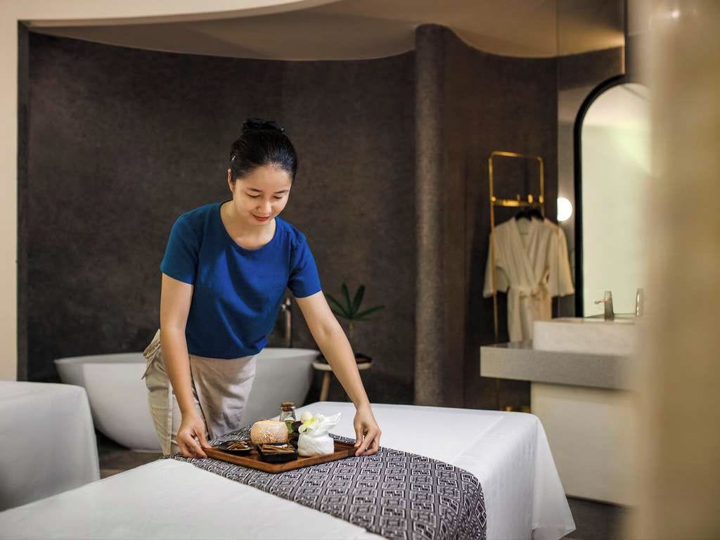 A person setting up a tray with amenities on a bed in a modern room with a large mirror and a sink.