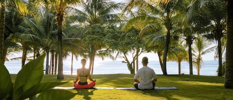 Dos personas meditan en un césped con palmeras y vista al mar.