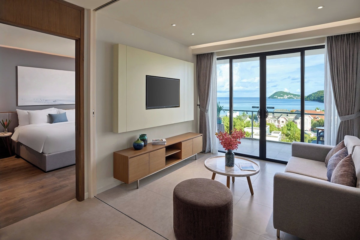 Quarto de hotel moderno com cama, TV fixada na parede, sofá, mesa de centro e vista para o mar.