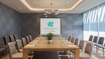 Un salón de conferencias con una larga mesa, sillas, una pantalla de proyector y una planta en maceta.
