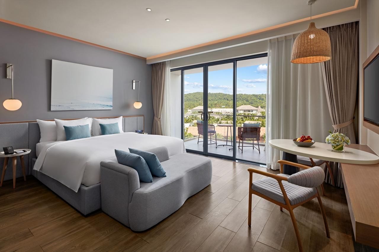 A modern hotel room with a large bed, a sofa, a dining table, and a view of the outdoors.