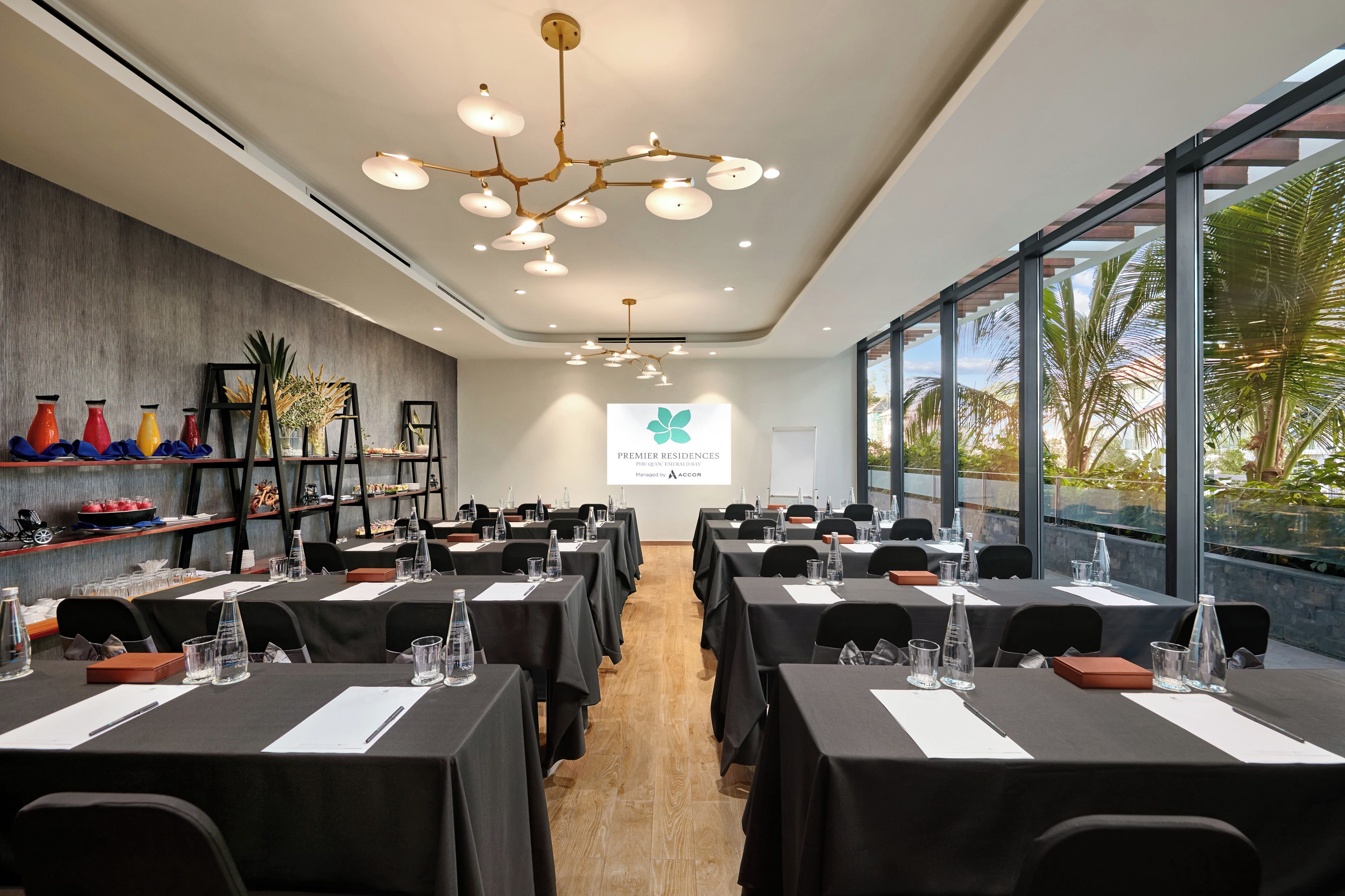 Un salón de conferencias con mesas dispuestas para reuniones, una pantalla de proyector y vistas a palmeras a través de amplios ventanales.