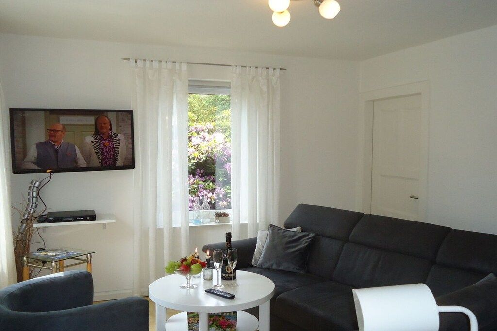Una sala de estar con un sofá negro, una mesa de centro blanca con un control remoto, una botella y vasos. Hay un televisor en la pared y una ventana con cortinas.