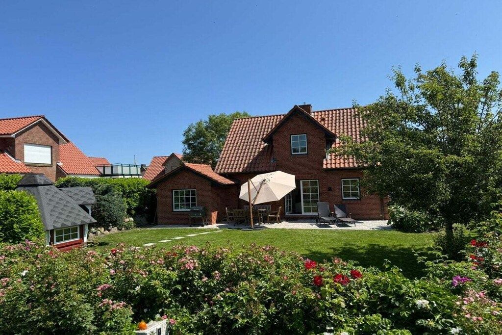 Una casa de dos pisos con techo de tejas rojas, un patio con mobiliario exterior y un jardín bien cuidado con diversas plantas y flores.