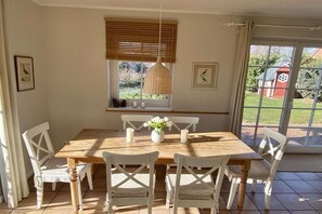 Un comedor con una mesa de madera, sillas blancas, una lámpara colgante y una ventana con persianas.