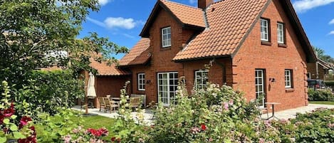 A two-story brick house with a sloped roof, a patio with outdoor furniture, and a garden with flowering plants.