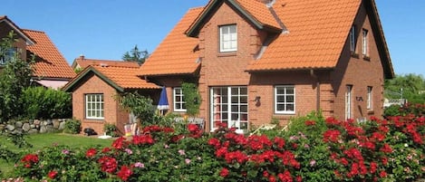 Une maison à deux étages, avec un toit recouvert de tuiles rouges, une façade en briques et un jardin agrémenté de fleurs rouges.