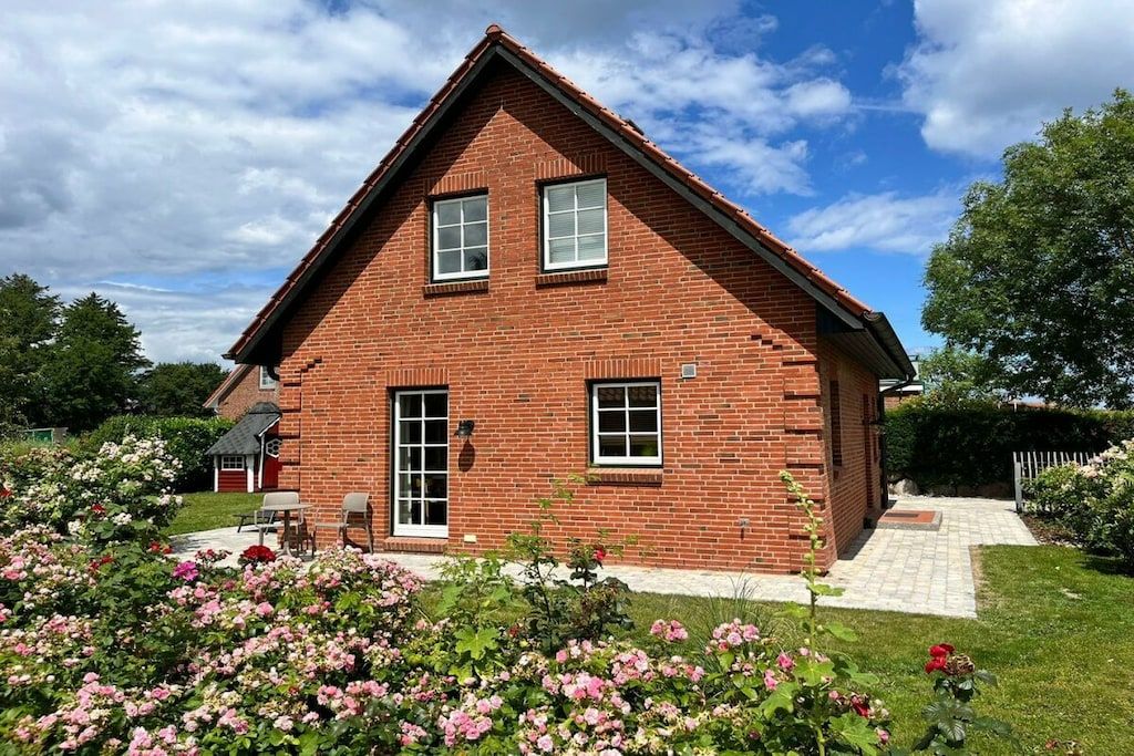 A two-story brick house with a sloped roof, a front porch, and a garden with pink flowers.