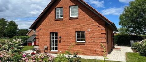 A two-story brick house with a sloped roof, a front porch, and a garden with pink flowers.