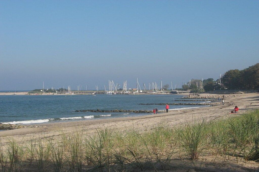 Uma praia com céu claro, mar calmo e turbinas eólicas ao longe.
