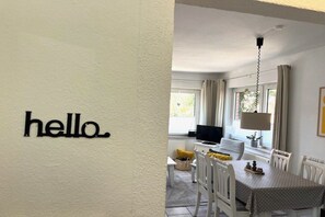 A room with a 'hello' sign, a dining table with chairs, a television, and a window with curtains. - Holiday apartment with 2 bedrooms (Fehmarn)