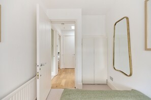 A hallway with a door leading to a room, a mirror on the wall, and a carpeted floor. - The Two-Bedroom Edit (London)