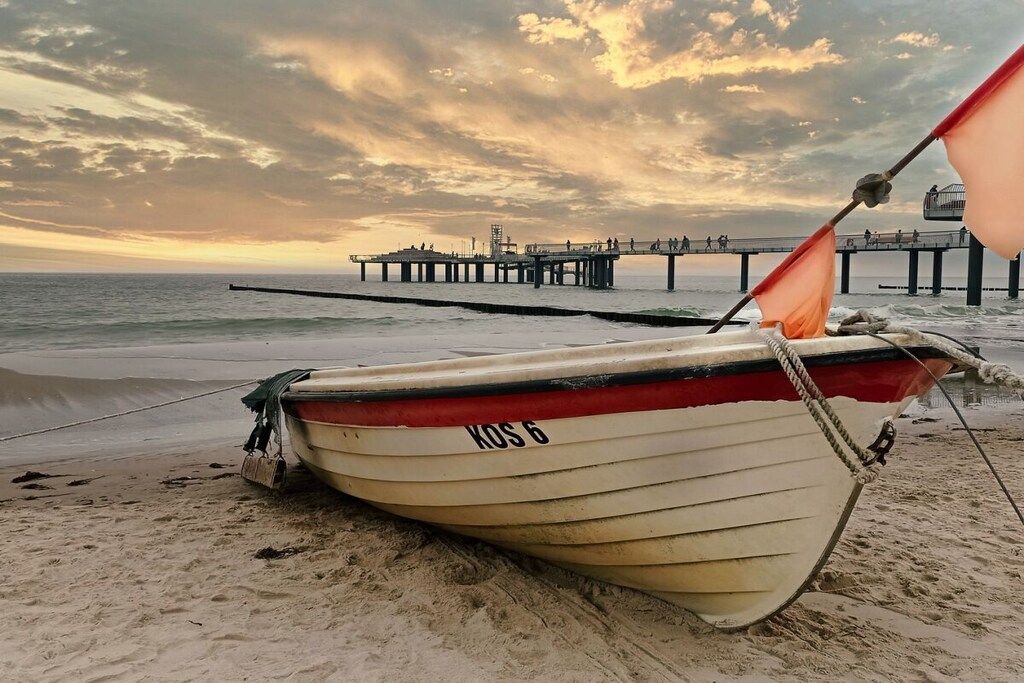 A boat with the identifier 'KOS 6' on a sandy beach, a pier in the background, and a cloudy sky.