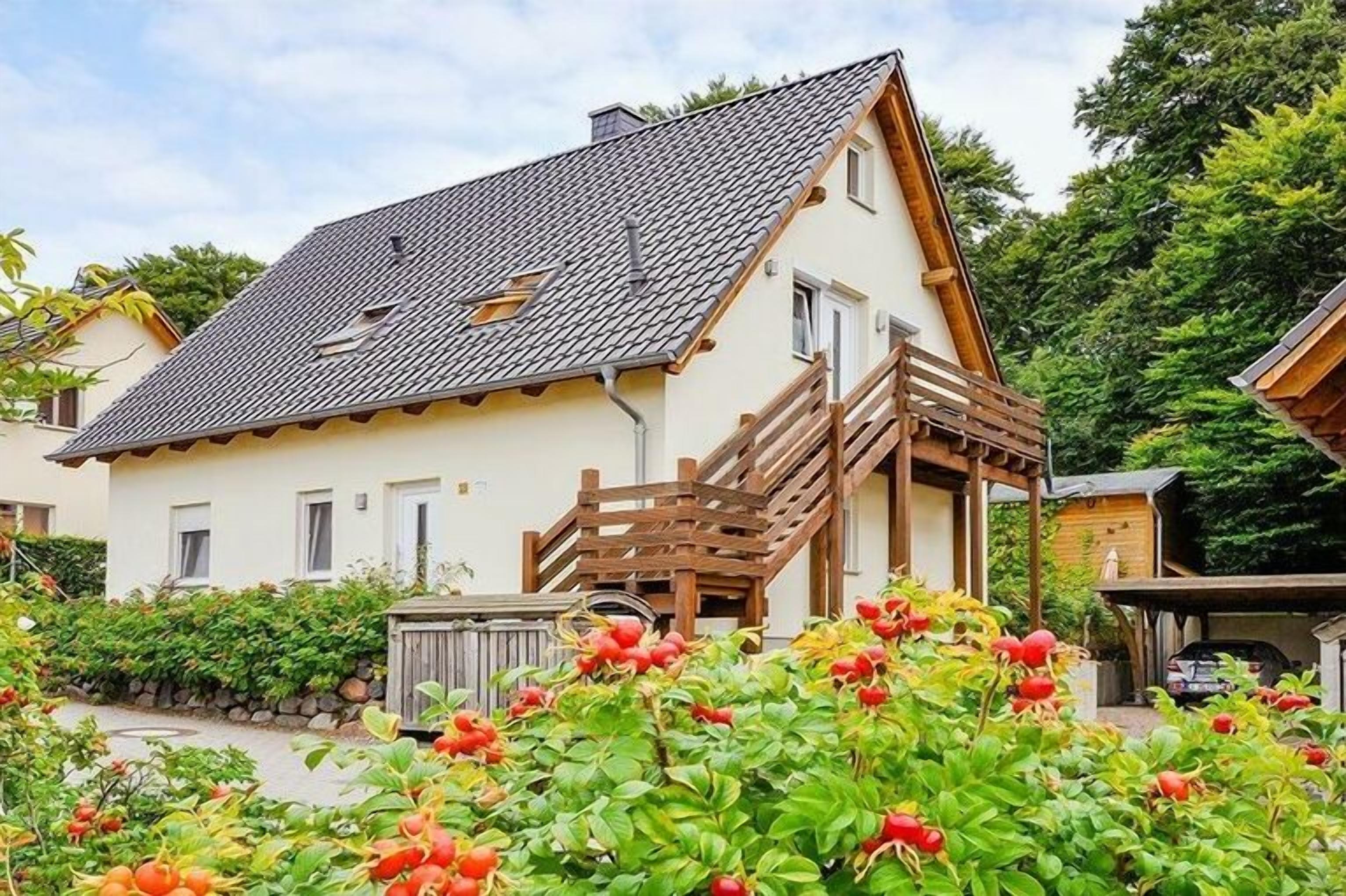 A two-story house with a balcony, surrounded by lush greenery and flowering plants.