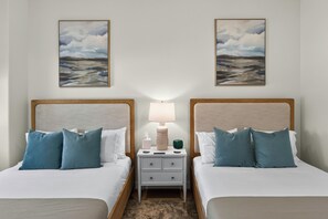 Quarto de hotel com duas camas, cada uma com cabeceira de madeira, roupa de cama branca e almofadas azuis. Há uma mesa de cabeceira com uma luminária, caixa de lenços e controle remoto entre as camas. Duas pinturas emolduradas de paisagens estão penduradas na parede acima das camas.