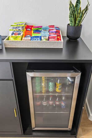 A snack tray with various popcorn and candy bars, a potted plant, and a mini fridge stocked with beverages. - Prime Location | New Luxury House | Walk to AT&T (Arlington)
