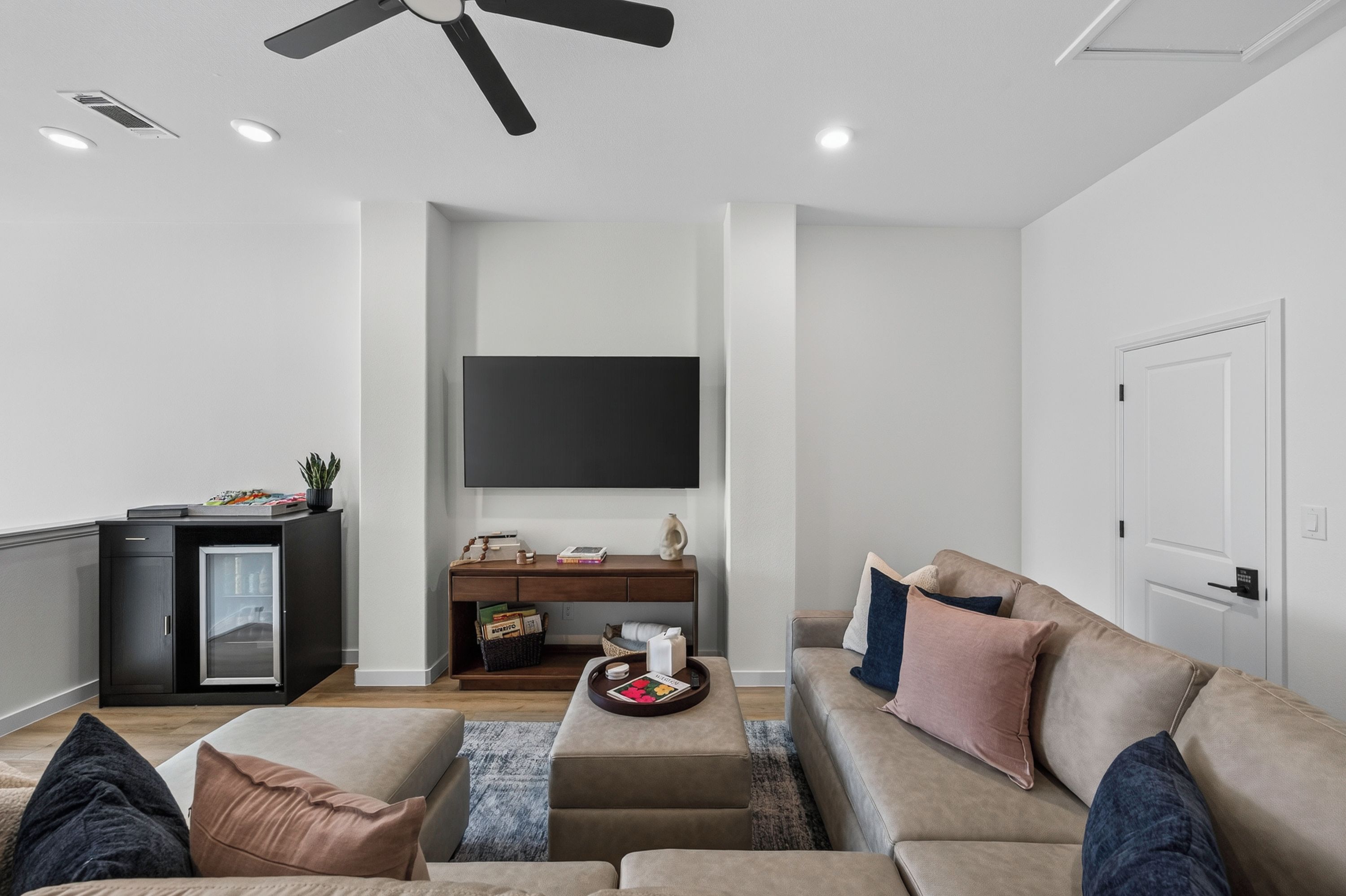 A modern living room with a flat-screen TV mounted on the wall, a dark wooden desk, a beige sofa, and a small coffee table.