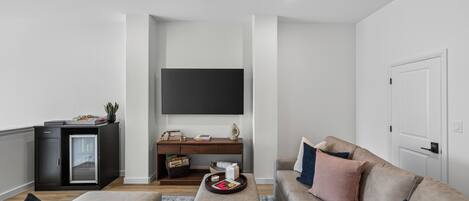 A modern living room with a flat-screen TV mounted on the wall, a dark wooden desk, a beige sofa, and a small coffee table.