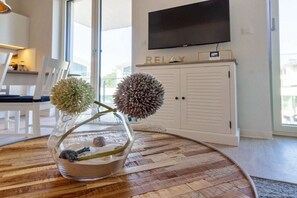 A modern living room with a glass vase containing two spherical objects, a flat-screen TV mounted above a cabinet, and a dining area with white chairs. - Holiday apartment in Glowe with swimming pool (Glowe)