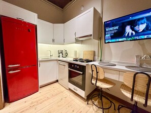 A compact kitchen with a red refrigerator, white cabinets, a built-in oven, and a flat-screen TV displaying a show. - Historic 2BRApt. with 2 Bathrooms, Turkish Bath & 3AC @Taksim (Istanbul)