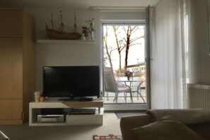 Ein Wohnzimmer mit einem Flachbildfernseher auf einem Schrank, einer Couch und einer Glastür, die zu einem Außenbereich mit Tisch und Stühlen führt.