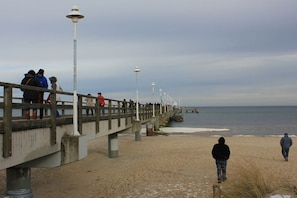 Eine Steg mit einem Gehweg zum Strand, ein sandiger Uferstreifen und das Meer im Hintergrund.