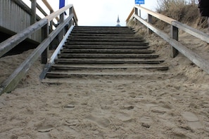 Eine Holztreppe führt zu einem Sandstrand, wobei ein blaues Schild auf einen Parkplatz hinweist.