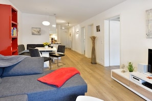 A modern living room with a grey sofa, a red bookshelf, a dining area with a table and chairs, and a TV on a white stand. - Holiday apartment with 2 bedrooms (Breege\/Juliusruh OT Juliusruh)