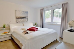 A neatly arranged bedroom with a bed, a bedside table, a lamp, a window with curtains, and a wall-mounted artwork. - Holiday apartment with 2 bedrooms (Breege\/Juliusruh OT Juliusruh)