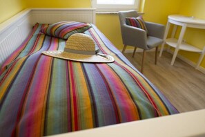 Un lit recouvert d’une couverture à rayures colorées, sur lequel est posé un chapeau de paille. La pièce contient également une chaise et une petite table.