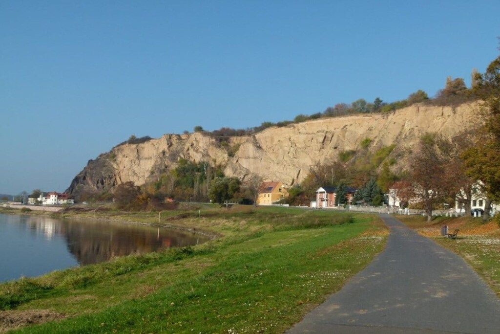 Un sentier le long de la rivière, avec une falaise, des maisons et un petit bâtiment.