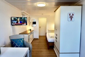 A compact room with a television mounted on the wall, a bed with a white mattress, a small table with a chair, and a wooden cabinet. - Holiday apartment with 1 bedroom (Wenningstedt-Braderup)