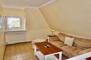 A living room with a beige sofa, a wooden coffee table, a white radiator, and a window with curtains. - Friesenhaus Carl Jensen Holiday Apartment 2 (Wenningstedt-Braderup)