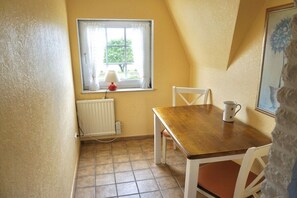 A small dining area with a table and chairs, a window with a view of greenery, and a radiator. - Friesenhaus Carl Jensen Holiday Apartment 2 (Wenningstedt-Braderup)