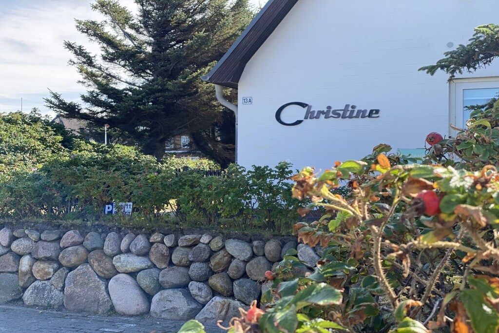 Un edificio con el nombre 'Christine' en su fachada, rodeado de vegetación y un muro de piedra.