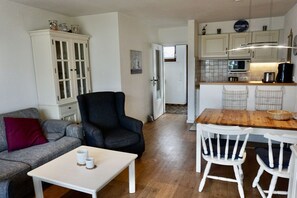 Ein kompakter Wohnbereich mit Küchenzeile, einer Essecke mit weißen Stühlen, einem grauen Sofa mit rotem Kissen und einem dunklen Sessel.