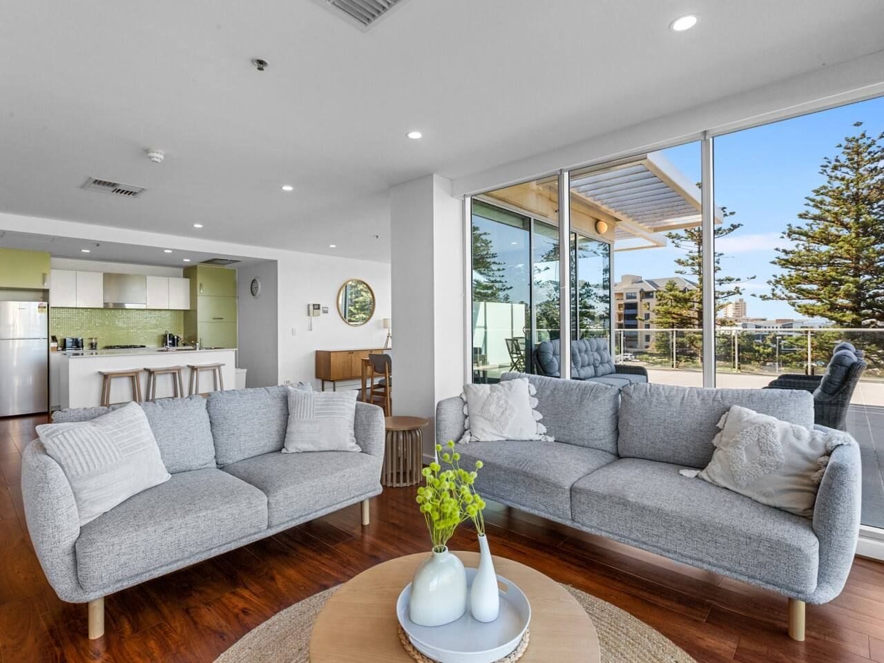 A modern living room with a grey sectional sofa, a coffee table with a vase, and a kitchen in the background.