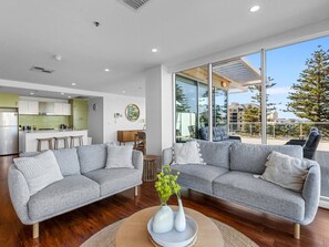 A modern living room with a grey sectional sofa, a coffee table with a vase, and a kitchen in the background. - Ocean Breeze - Stylish 2 Bedroom with terrace - 3-minute walk to beach (Glenelg)