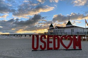 Ein Strand mit einem großen roten Schild, auf dem 'USEDOM' steht, und ein Gebäude mit zwei grünen Dächern im Hintergrund.