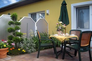 Eine Terrasse im Freien mit einem für zwei Personen gedecktem Tisch, umgeben von Blumentöpfen und einer gelben Wand.