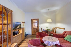 A furnished living room with a television, a wooden cabinet with glass doors, a dining table with chairs, and a red sofa. - House by the Forest Apartment 3 (Seebad Ahlbeck)