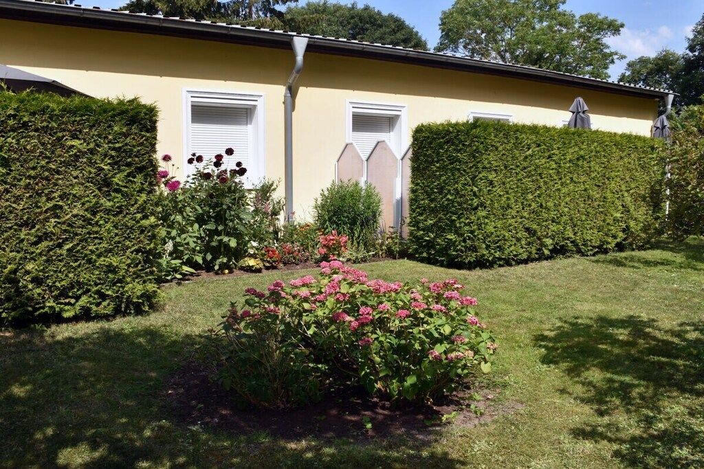 Un bâtiment jaune aux volets blancs, entouré de haies bien entretenues et de plantes en fleurs.
