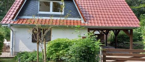 Ein kleines Haus mit rotem Ziegeldach, einer Holzveranda und einem eingezäunten Garten.