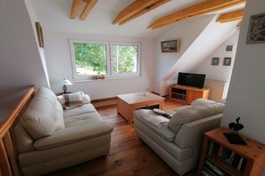 Ein gemütliches Wohnzimmer mit einer Couch, einem Couchtisch und einem Fernseher. Fenster bieten Blick auf Grünflächen außerhalb.