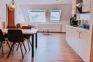 A modern kitchen with white cabinets, a dining area with a wooden table and chairs, and two windows with landscape views.