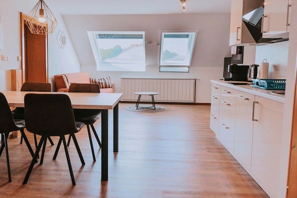 A modern kitchen with white cabinets, a dining area with a wooden table and chairs, and two windows with landscape views.