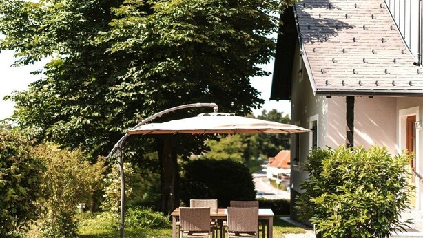 An outdoor patio with a table and chairs under a large umbrella, surrounded by greenery and a building.