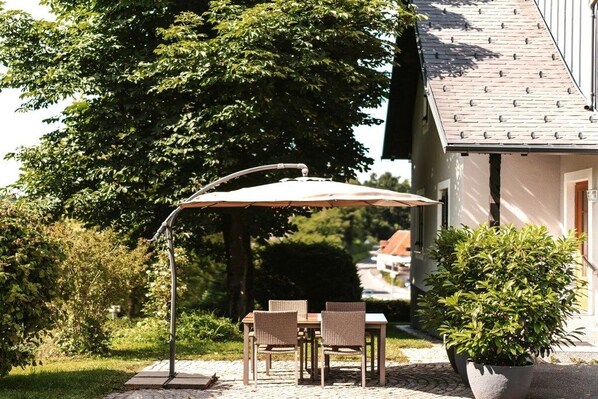 An outdoor patio with a table and chairs under a large umbrella, surrounded by greenery and a building.